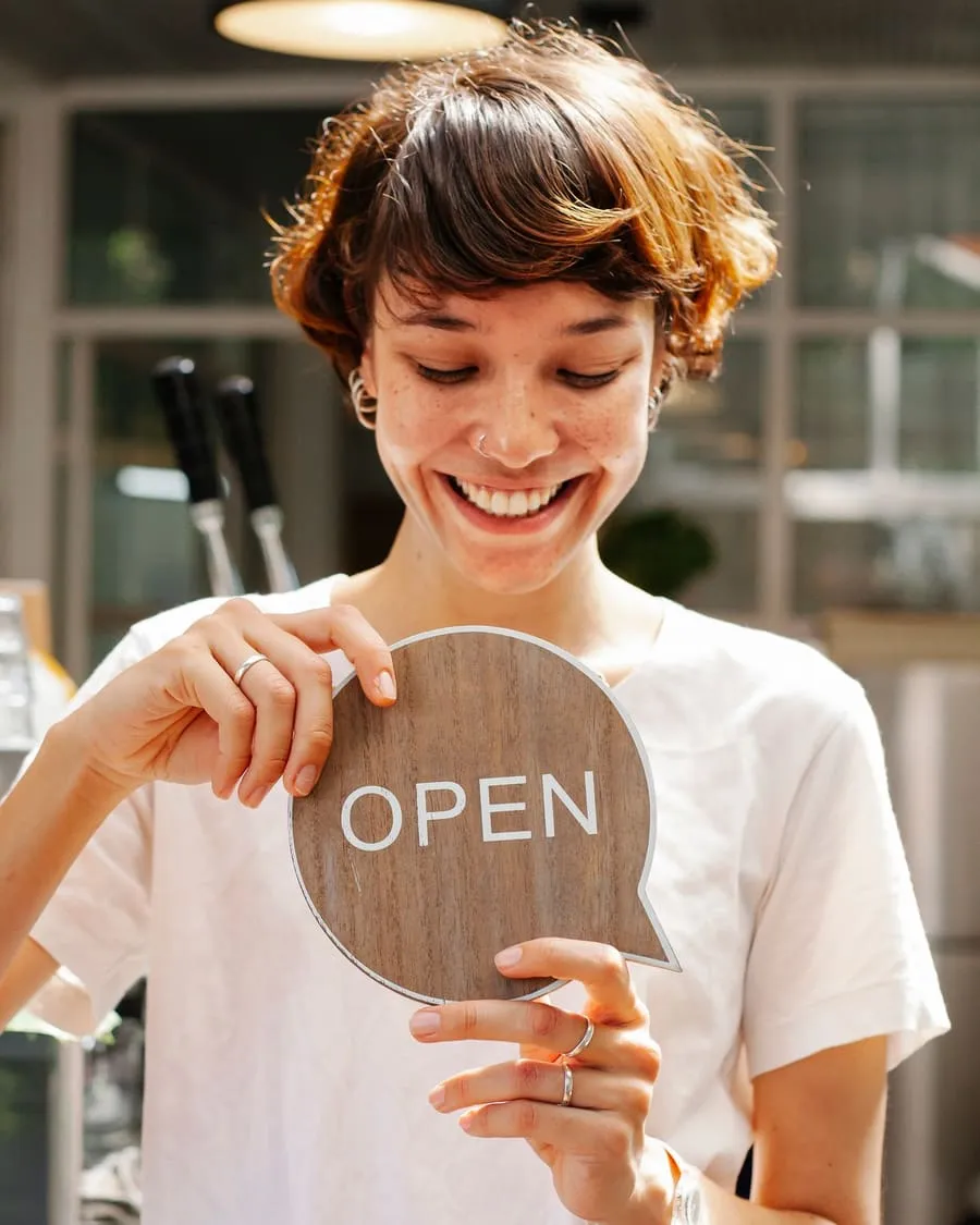 Atendente sorrindo e segurando uma placa de "aberto" na porta de uma cafeteria