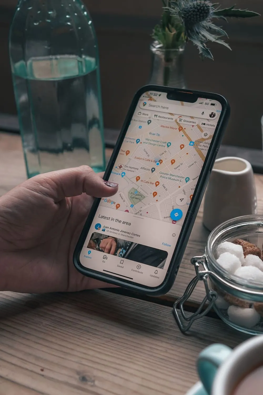 Mão segurando um celular com o mapa de lugares próximos aberto, sobre a mesa de um café